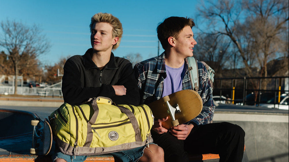 Two young men sitting on a bench with skateboards, one holding a yellow bag.